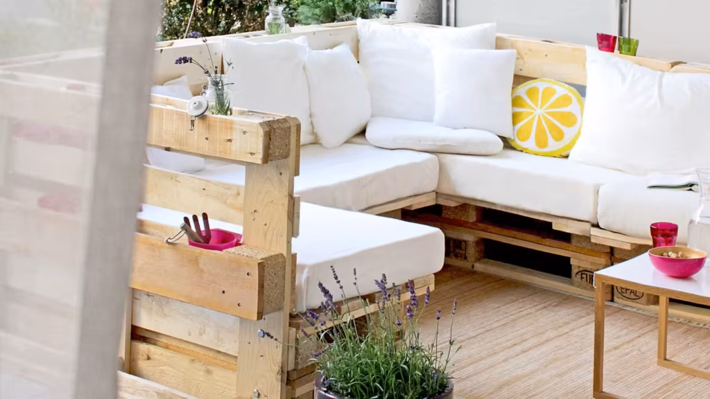 „Gemütliche Sitzecke aus hellen Palettenmöbeln mit weißen Polstern und vielen Kissen. Die Lounge steht auf einer Terrasse und ist dekoriert mit Pflanzen, bunten Gläsern und einem Zitronen-Kissen für einen frischen, einladenden Look.“