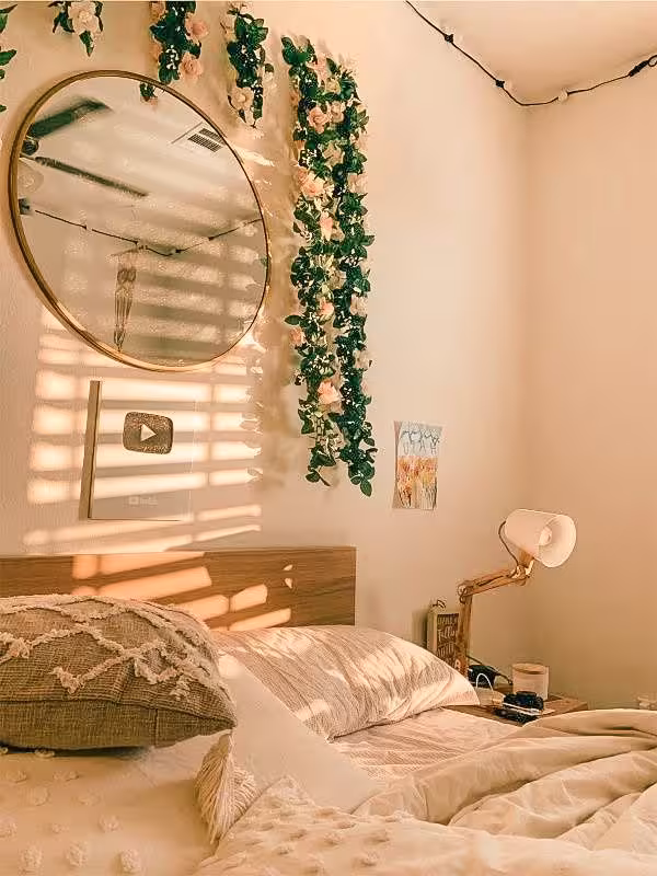„Gemütlich dekoriertes Schlafzimmer mit einem großen Bett, einem runden Spiegel über dem Kopfteil, herabhängenden Blumenranken und sanftem Sonnenlicht, das durch Jalousien fällt. Eine warme, ästhetische Atmosphäre im Boho-Stil.“