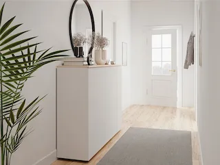 Heller, modern eingerichteter Flur mit weißer Kommode auf der linken Seite. Auf der Kommode stehen dekorative Vasen mit Trockenblumen und ein rundes Spiegelobjekt. Der Boden ist aus hellem Holz, darauf liegt ein grauer Teppichläufer. Im Hintergrund befindet sich eine weiße Tür mit Glaseinsatz, durch die Tageslicht einfällt. Eine grüne Zimmerpflanze steht links im Vordergrund.